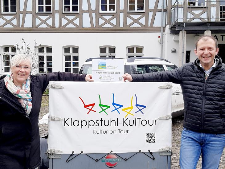 Bürgermeister Marco Trautner übergibt Förderschild an eine Frau vor dem Anhänger der Klappstuhl-KulTour